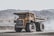 A large yellow dump truck is situated in an expansive quarry or mining site, surrounded by rocky, mountainous terrain. The truck is stationary, with dust or smoke around it, indicating recent movement.