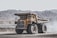 A large yellow dump truck is situated in an expansive quarry or mining site, surrounded by rocky, mountainous terrain. The truck is stationary, with dust or smoke around it, indicating recent movement.