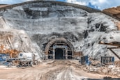 A large tunnel entrance carved into a rocky hillside is surrounded by scaffolding and construction equipment. The ground is covered in dirt and mud, indicating an active construction site. Several machines and vehicles, including a water truck, are scattered around the area. The earthy tones of the rocks contrast with the bright blue sky above.