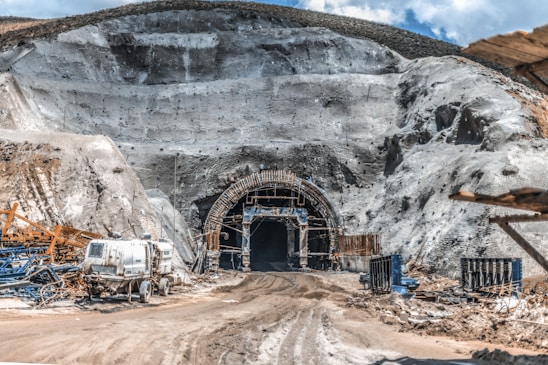 A large tunnel entrance carved into a rocky hillside is surrounded by scaffolding and construction equipment. The ground is covered in dirt and mud, indicating an active construction site. Several machines and vehicles, including a water truck, are scattered around the area. The earthy tones of the rocks contrast with the bright blue sky above.