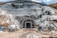A large tunnel entrance carved into a rocky hillside is surrounded by scaffolding and construction equipment. The ground is covered in dirt and mud, indicating an active construction site. Several machines and vehicles, including a water truck, are scattered around the area. The earthy tones of the rocks contrast with the bright blue sky above.
