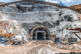 A large tunnel entrance carved into a rocky hillside is surrounded by scaffolding and construction equipment. The ground is covered in dirt and mud, indicating an active construction site. Several machines and vehicles, including a water truck, are scattered around the area. The earthy tones of the rocks contrast with the bright blue sky above.