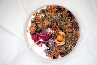 A bowl filled with yogurt is topped with a variety of colorful ingredients, including granola, blueberries, raspberries, pumpkin seeds, and gooseberries. The mixture also contains chia seeds and is drizzled with a berry sauce. A pink spoon rests on the edge of the bowl.