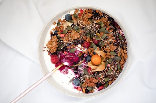 A bowl filled with yogurt is topped with a variety of colorful ingredients, including granola, blueberries, raspberries, pumpkin seeds, and gooseberries. The mixture also contains chia seeds and is drizzled with a berry sauce. A pink spoon rests on the edge of the bowl.
