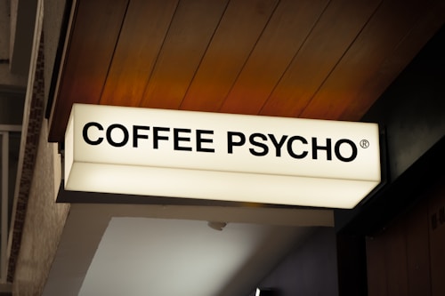 A rectangular lightbox sign with the bold, black text 'COFFEE PSYCHO' in uppercase letters. The sign is mounted on a building exterior, featuring a wooden paneled ceiling. The overall setting is urban, possibly suggesting a coffee shop or cafe.
