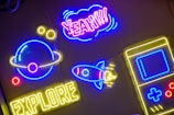 Brightly colored neon signs are mounted against a dark background. A neon planet with rings is in blue and yellow, accompanied by small yellow circles. The word 'EXPLORER' is lit up in yellow at the bottom. Above it, a neon rocket with blue and red accents points upwards. The word 'YEAH!!!' is prominently displayed in purple and pink, encased in a cloud-shaped outline. A yellow and blue outline of a handheld gaming device is also visible.