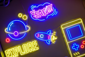 Brightly colored neon signs are mounted against a dark background. A neon planet with rings is in blue and yellow, accompanied by small yellow circles. The word 'EXPLORER' is lit up in yellow at the bottom. Above it, a neon rocket with blue and red accents points upwards. The word 'YEAH!!!' is prominently displayed in purple and pink, encased in a cloud-shaped outline. A yellow and blue outline of a handheld gaming device is also visible.