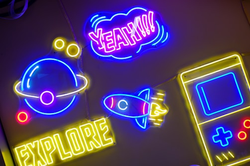 Brightly colored neon signs are mounted against a dark background. A neon planet with rings is in blue and yellow, accompanied by small yellow circles. The word 'EXPLORER' is lit up in yellow at the bottom. Above it, a neon rocket with blue and red accents points upwards. The word 'YEAH!!!' is prominently displayed in purple and pink, encased in a cloud-shaped outline. A yellow and blue outline of a handheld gaming device is also visible.