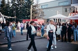 A bustling outdoor gathering with a diverse group of people walking and socializing. The setting includes trees, a modern building, and red shipping containers. Strung lights and large umbrellas create a festive atmosphere, hinting at an event or fair. The crowd is engaged in various activities like walking, talking, and gathering around tables.
