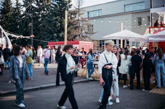 A bustling outdoor gathering with a diverse group of people walking and socializing. The setting includes trees, a modern building, and red shipping containers. Strung lights and large umbrellas create a festive atmosphere, hinting at an event or fair. The crowd is engaged in various activities like walking, talking, and gathering around tables.
