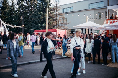 A bustling outdoor gathering with a diverse group of people walking and socializing. The setting includes trees, a modern building, and red shipping containers. Strung lights and large umbrellas create a festive atmosphere, hinting at an event or fair. The crowd is engaged in various activities like walking, talking, and gathering around tables.