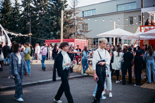 A bustling outdoor gathering with a diverse group of people walking and socializing. The setting includes trees, a modern building, and red shipping containers. Strung lights and large umbrellas create a festive atmosphere, hinting at an event or fair. The crowd is engaged in various activities like walking, talking, and gathering around tables.
