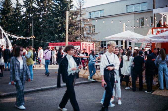 A bustling outdoor gathering with a diverse group of people walking and socializing. The setting includes trees, a modern building, and red shipping containers. Strung lights and large umbrellas create a festive atmosphere, hinting at an event or fair. The crowd is engaged in various activities like walking, talking, and gathering around tables.