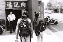Two people with backpacks walk down a city sidewalk in an urban setting. A man in a white shirt stands in front of a store with signage in an Asian script. Scooters are parked by the roadside under a sunlit sky, while trees and urban structures are visible in the background.