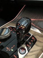 A close-up of a car's center console featuring a crystal gear shifter, a rotary dial, and several illuminated buttons. The panel displays indicators for various driving modes and the start/stop button is prominently visible. The console has a glossy black finish with red and green lighting accents.