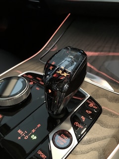 A close-up of a car's center console featuring a crystal gear shifter, a rotary dial, and several illuminated buttons. The panel displays indicators for various driving modes and the start/stop button is prominently visible. The console has a glossy black finish with red and green lighting accents.