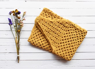 A hand-knitted yellow scarf with a textured, open weave pattern is placed on a white wooden surface next to a small bundle of dried wildflowers, including yellow and purple blooms.