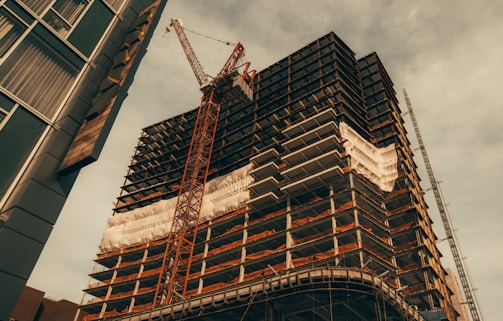 A tall building under construction is depicted with metal scaffolding and cranes. The structure is partially covered with a white protective wrap. The sky above is cloudy and the angle of the shot gives a dramatic perspective.