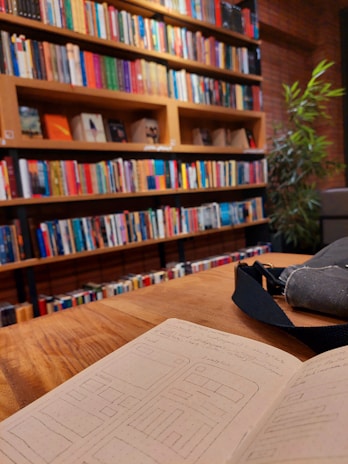 A cozy library scene with shelves filled with colorful books arranged neatly. In the foreground, an open notebook with sketches and writings is placed on a wooden table. Adjacent to the notebook is a gray bag. A leafy green plant is visible in the background, adding a touch of nature to the setting.