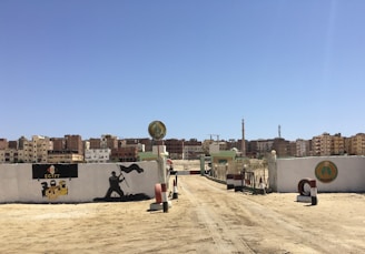 A sandy dirt road leads to a gated entrance with painted walls on either side. The wall on the left depicts a silhouette of a soldier holding a flag and text mentioning 'EGYPT'. There are residential buildings in the distance under a clear blue sky.