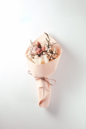 A delicate bouquet of flowers wrapped in light pink paper, featuring a mix of roses and small white blossoms with some greenery. A ribbon tied around the wrapping adds a touch of elegance.