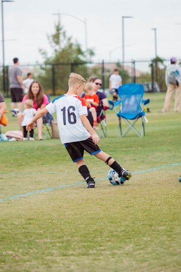 A child wearing a soccer jersey with the number 16 is actively playing with a soccer ball on a grass field. Spectators, including adults and children, are seated in the background on folding chairs, observing the game. The scene is set outdoors with a fence lining the edge of the field and trees in the distance.