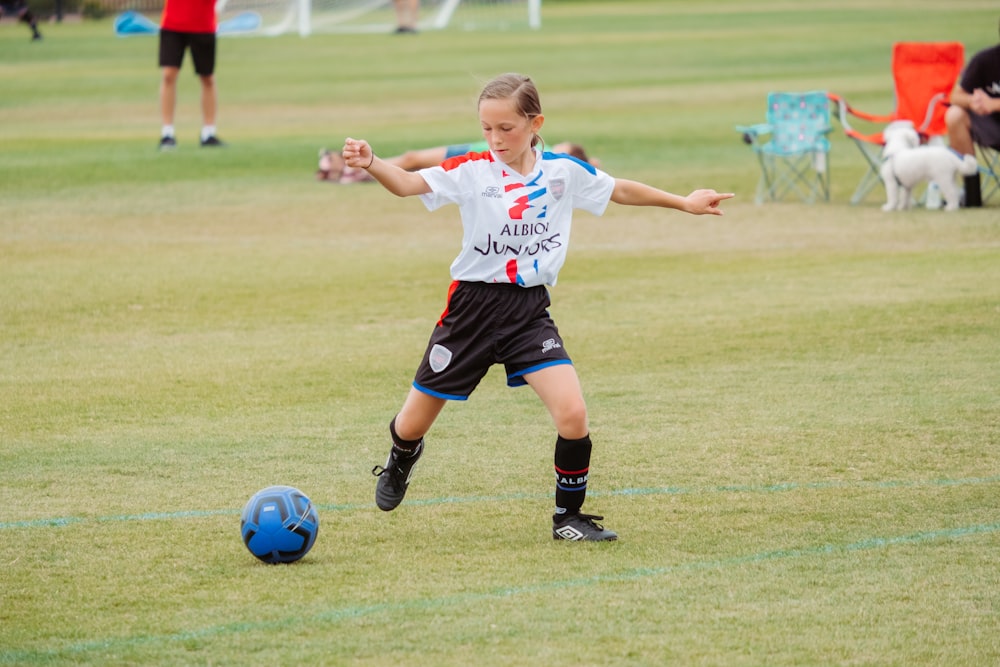 흰색 셔츠와 검은 반바지를 입은 소녀는 낮 동안 푸른 잔디밭에서 축구를 하고 있다