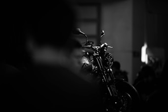 A motorcycle is highlighted in a dimly lit setting, with its silhouette and details sharply defined against a blurred, dark background. The focus is on the front of the bike, emphasizing its sleek design and metallic elements.