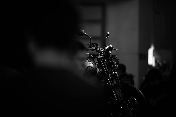 A motorcycle is highlighted in a dimly lit setting, with its silhouette and details sharply defined against a blurred, dark background. The focus is on the front of the bike, emphasizing its sleek design and metallic elements.