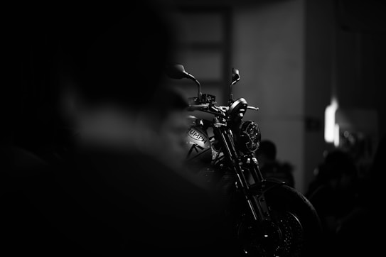 A motorcycle is highlighted in a dimly lit setting, with its silhouette and details sharply defined against a blurred, dark background. The focus is on the front of the bike, emphasizing its sleek design and metallic elements.