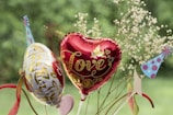 A red heart-shaped balloon with the word 'Love' and decorated with gold patterns is accompanied by a white balloon with gold and red designs. There are also small delicate flowers in the background and a couple of colorful, polka-dotted party hats.