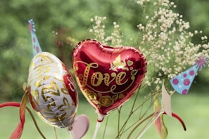 A red heart-shaped balloon with the word 'Love' and decorated with gold patterns is accompanied by a white balloon with gold and red designs. There are also small delicate flowers in the background and a couple of colorful, polka-dotted party hats.