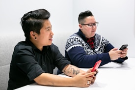 Two people are sitting next to each other at a table, both holding smartphones. One person is wearing a black short-sleeve shirt with tattoos on their arm, while the other is wearing a blue and white patterned sweater over a plaid shirt. They appear to be focused on their devices in a well-lit environment.