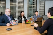 A group of four individuals is seated around a wooden conference table in a modern office setting. Laptops with decorated covers are in front of three people, while two large windows reveal tall office buildings in the background. The group appears to be engaged in a conversation, and they are smiling, creating a welcoming atmosphere.