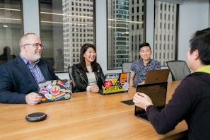 A group of four individuals is seated around a wooden conference table in a modern office setting. Laptops with decorated covers are in front of three people, while two large windows reveal tall office buildings in the background. The group appears to be engaged in a conversation, and they are smiling, creating a welcoming atmosphere.