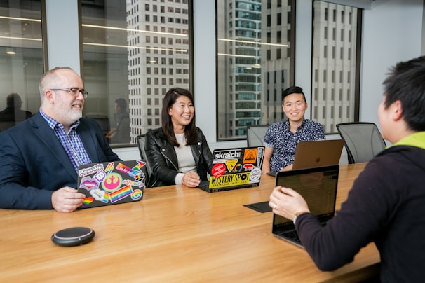A group of four individuals is seated around a wooden conference table in a modern office setting. Laptops with decorated covers are in front of three people, while two large windows reveal tall office buildings in the background. The group appears to be engaged in a conversation, and they are smiling, creating a welcoming atmosphere.