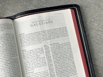 An open Bible lies on a gray fabric surface, displaying the pages from 'The Letter of Paul to the Galatians'. The text is in a double-column format with chapter headings and verses clearly visible. The edges of the pages are a reddish color, and the book has a black cover with golden detailing along the edge.