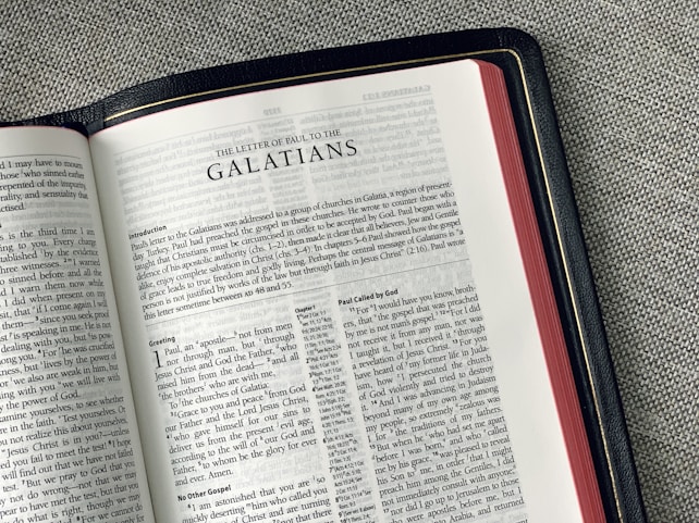 An open Bible lies on a gray fabric surface, displaying the pages from 'The Letter of Paul to the Galatians'. The text is in a double-column format with chapter headings and verses clearly visible. The edges of the pages are a reddish color, and the book has a black cover with golden detailing along the edge.