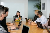 A group of six people are seated around a wooden conference table, engaged in a team meeting. Each person has a laptop in front of them, with the central figure speaking and others listening attentively. The room has a modern appearance, with a whiteboard on the wall and a potted plant in the corner.