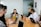 A group of six people are seated around a wooden conference table, engaged in a team meeting. Each person has a laptop in front of them, with the central figure speaking and others listening attentively. The room has a modern appearance, with a whiteboard on the wall and a potted plant in the corner.