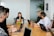 A group of six people are seated around a wooden conference table, engaged in a team meeting. Each person has a laptop in front of them, with the central figure speaking and others listening attentively. The room has a modern appearance, with a whiteboard on the wall and a potted plant in the corner.