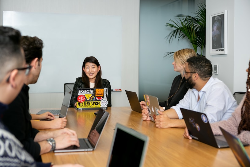 A group of six people are seated around a wooden conference table, engaged in a team meeting. Each person has a laptop in front of them, with the central figure speaking and others listening attentively. The room has a modern appearance, with a whiteboard on the wall and a potted plant in the corner.
