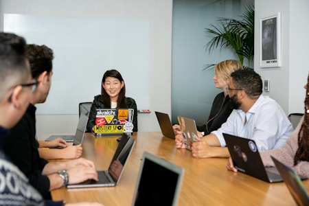 A group of six people are seated around a wooden conference table, engaged in a team meeting. Each person has a laptop in front of them, with the central figure speaking and others listening attentively. The room has a modern appearance, with a whiteboard on the wall and a potted plant in the corner.
