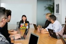 A group of six people are seated around a wooden conference table, engaged in a team meeting. Each person has a laptop in front of them, with the central figure speaking and others listening attentively. The room has a modern appearance, with a whiteboard on the wall and a potted plant in the corner.