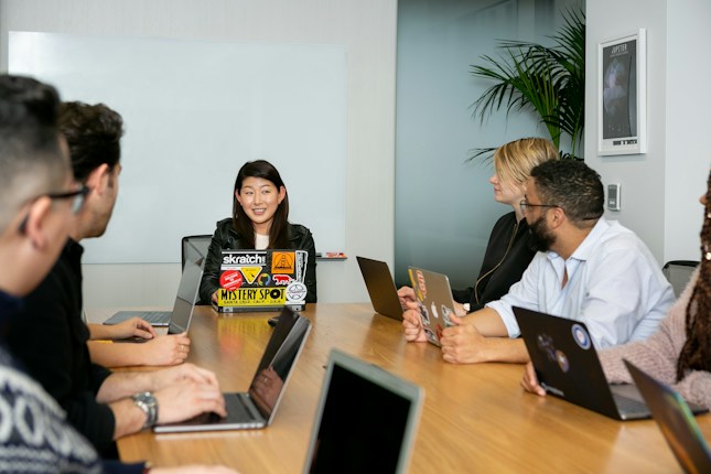 A group of six people are seated around a wooden conference table, engaged in a team meeting. Each person has a laptop in front of them, with the central figure speaking and others listening attentively. The room has a modern appearance, with a whiteboard on the wall and a potted plant in the corner.