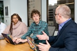 Three people are seated at a conference table with laptops in front of them. They appear to be engaged in a discussion or meeting. The person on the left has long braids and glasses, wearing a cozy light brown sweater. The person in the middle is wearing a green checked shirt. The person on the right is an older man with glasses, a beard, and a blue blazer, gesturing with his hands while speaking.