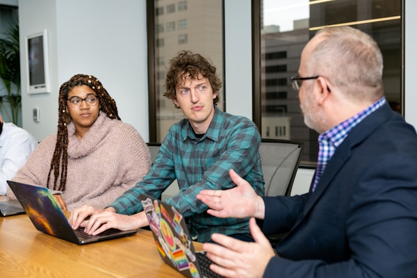 Three people are seated at a conference table with laptops in front of them. They appear to be engaged in a discussion or meeting. The person on the left has long braids and glasses, wearing a cozy light brown sweater. The person in the middle is wearing a green checked shirt. The person on the right is an older man with glasses, a beard, and a blue blazer, gesturing with his hands while speaking.