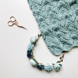 A light blue knitted fabric with a wavy, textured pattern is partially shown. Adjacent to it, there is a pair of small, ornate embroidery scissors with a handle shaped like a bird. Below, a floral garland made of artificial flowers in soft blues and greens rests gently on the surface.
