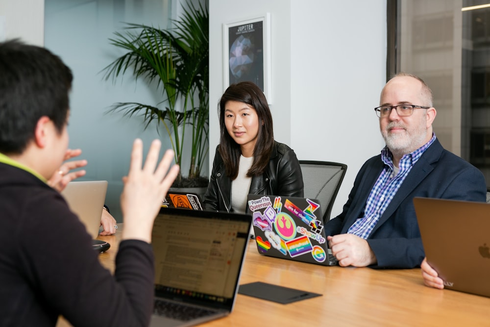 boardroom meeting where woman in black leather jacket and man in blue checkered shirt, suit jacket and glasses, pay attention to person talking with their hands wearing black long sleeve shirt