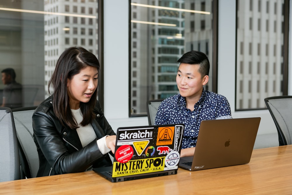 woman in black leather jacket points to laptop covered in stickers, sitting beside person in blue shirt with silver laptop